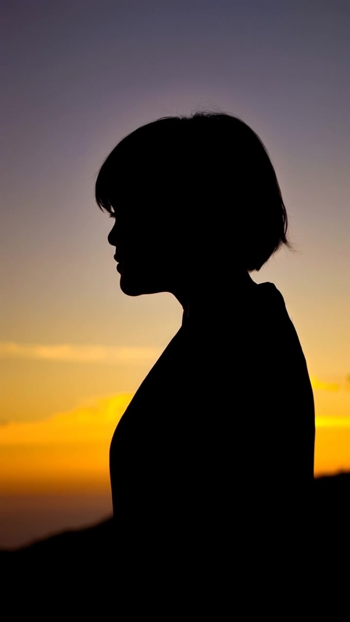 Subject: A person standing on a hillside at sunset. Subject movement: The figure shifts slightly as the wind gently sways their hair, capturing a moment of determination while gazing into the distance. Scene: A picturesque hillside with rolling grasses bathed in warm, golden light from the setting sun, casting long shadows. Lighting: Sunset lighting creating a warm and inviting atmosphere. Motion: Camera slowly pushes in to capture the contemplative expression, transitioning from the expansive hillside backdrop to a close-up view of the person, highlighting the emotion in their gaze.