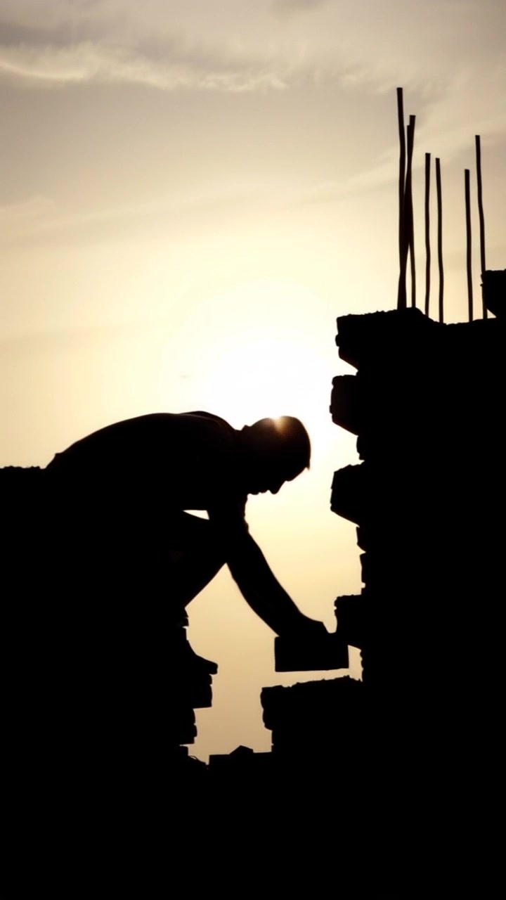 Subject: A construction worker laying bricks. Subject movement: The worker moves rhythmically, placing each brick with care under a gray sky. Scene: A bustling construction site surrounded by machinery and raw materials. Style: Overcast lighting enhancing the somber mood of a work-in-progress environment. Motion: The camera does a steady push-in, capturing the focused expression of the worker as he sets another brick in place, with a slight pan revealing the surrounding site.