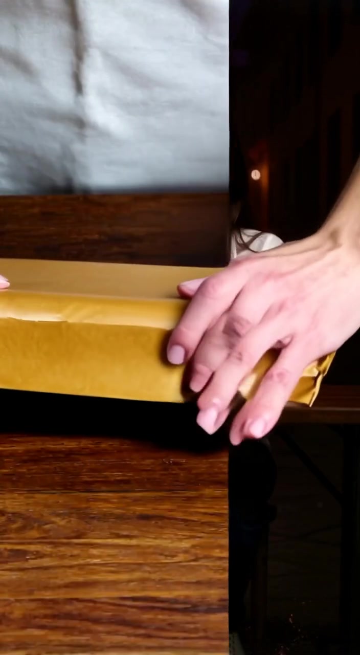 The woman gently unwraps the brown paper, revealing the small parcel on the wooden table.
