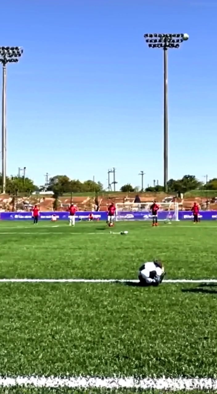 The boy skillfully dribbles the ball, maneuvering past players on an expansive soccer field under a clear blue sky.