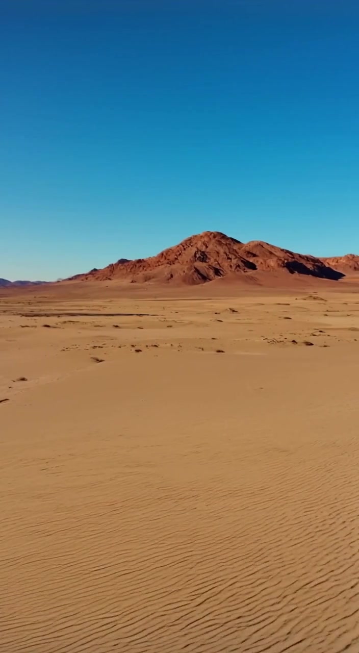A gentle breeze stirs the sand while distant mountains loom against a clear blue sky in the vast desert.