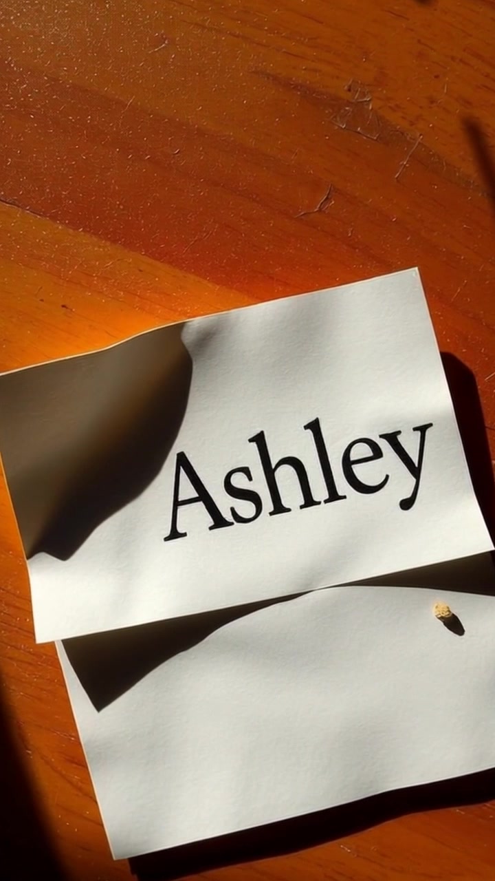 Scene: A close-up view of a note with the name 'Ashley' partially unrolled on a rustic wooden surface. Style: Soft, natural lighting highlighting the texture of the wood and the paper. Motion: Camera gently pushes in, focusing on the detail of the note while the surrounding environment softens into a dreamy bokeh.