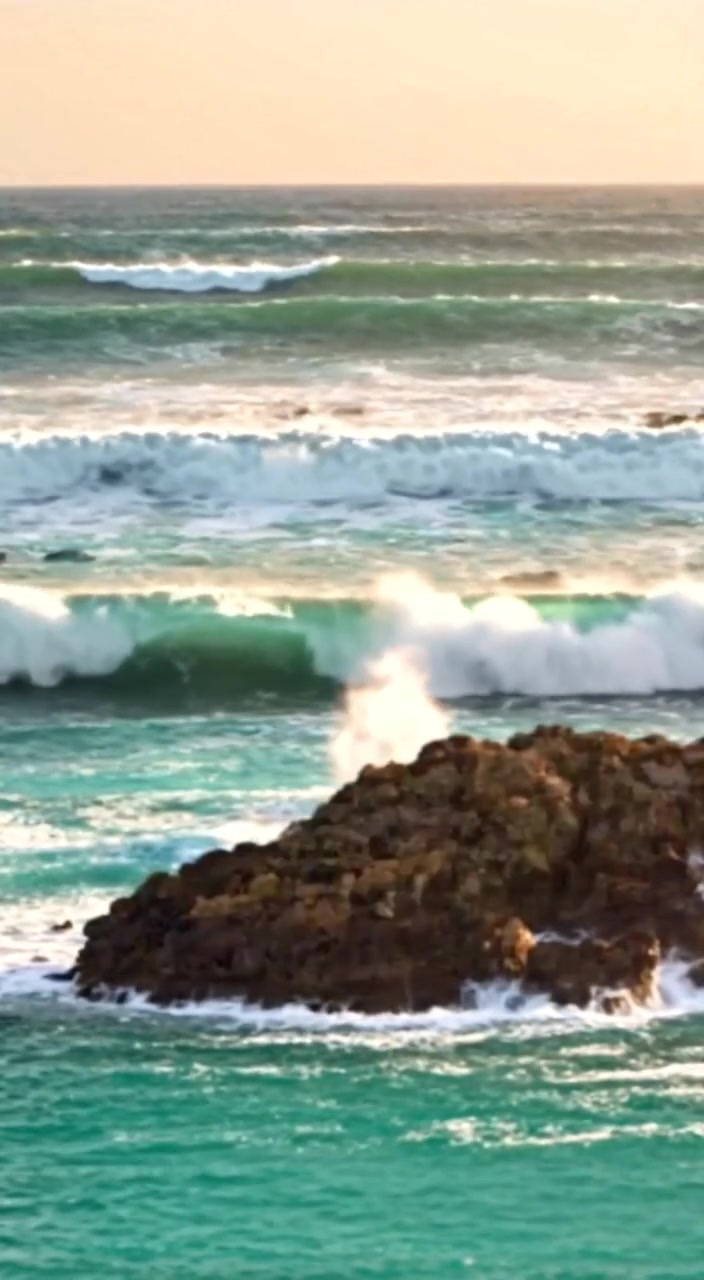 The woman stands on a rocky cliff as waves crash below, her hair gently blowing in the fierce wind.