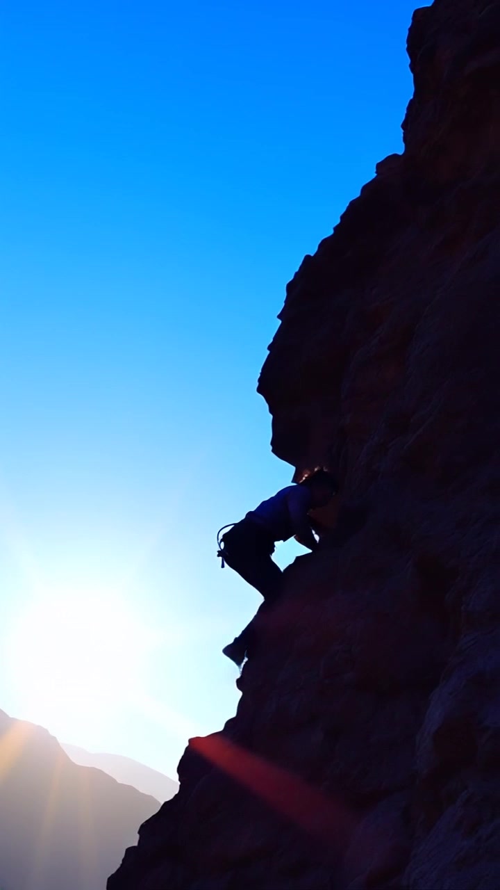 Subject: A determined climber ascending a steep mountain path. Subject movement: The climber takes deliberate, powerful steps, grasping for handholds and pushing upward with resolve. Scene: Rugged mountain landscape with rocky terrain and a blue sky above. Style: Bright daylight illuminating the rocks, showcasing contrasts between shadows and sunlit areas. Motion: The camera follows the climber closely from a slightly low angle, panning upward to capture the ascent while emphasizing the scale of the mountain.
