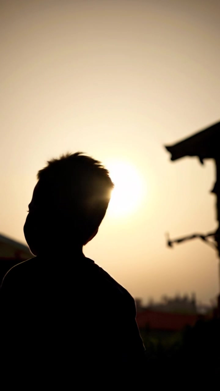 Subject: A small boy standing in the village, looking around with a curious expression. Subject movement: The boy takes a few hesitant steps forward, exploring his surroundings. Scene: A vibrant village setting, with colorful houses and lush greenery in the background. Style: Morning light gently illuminating the scene, creating a warm and inviting atmosphere. Motion: The camera slowly pushes in closer to capture the boy's fascinated gaze, then pans to reveal the bustling life of the village around him.