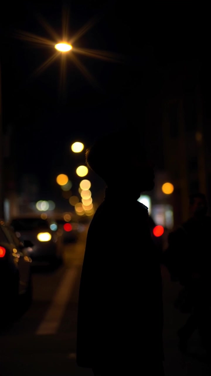 Subject: Isaiah walking alone. Subject movement: With determination, Isaiah strides purposefully down a dimly lit city street. Scene: A dark city scene illuminated by glowing streetlights, casting long shadows and creating a moody atmosphere. Style: The lighting is dramatic and atmospheric, with contrast between light and shadow adding depth. Motion: The camera follows Isaiah closely, offering a handheld tracking shot as he moves forward, panning smoothly to capture the surrounding cityscape.
