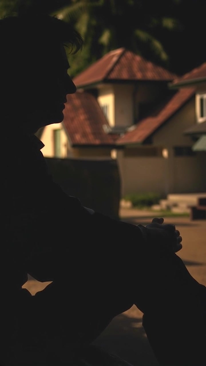 Subject: A person sitting on a bench, deep in thought. Subject movement: The person shifts slightly, adjusting their position as they gaze off into the distance. Scene: A tranquil village landscape, complete with lush green fields and quaint houses in the background. Style: Soft, ambient morning light illuminating the scene with a serene glow. Motion: The camera gently pushes in from a distance, drawing closer to the individual, capturing the subtle movements and contemplative atmosphere.