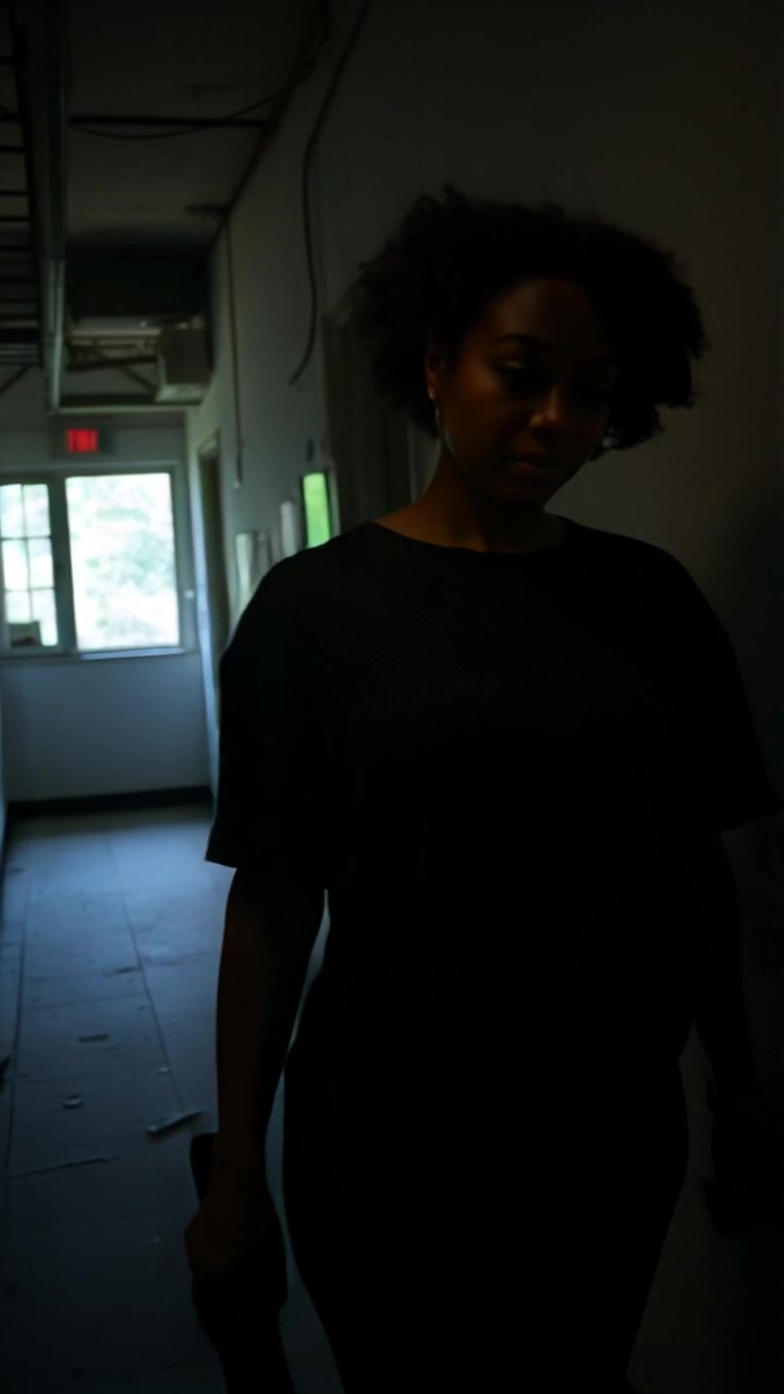 Subject: Nia moving cautiously through a dark hallway. Subject movement: Nia steps quietly, her flashlight beam cutting through the darkness, revealing her surroundings. Scene: An eerie, dimly lit hallway with shadows lurking in the corners, creating a tense atmosphere. Lighting: Low ambient light with sharp contrasts from the flashlight. Motion: Start with a close-up on Nia’s face as she focuses ahead, then pull back to reveal her moving further down the corridor, the beam of light sweeping across the walls.