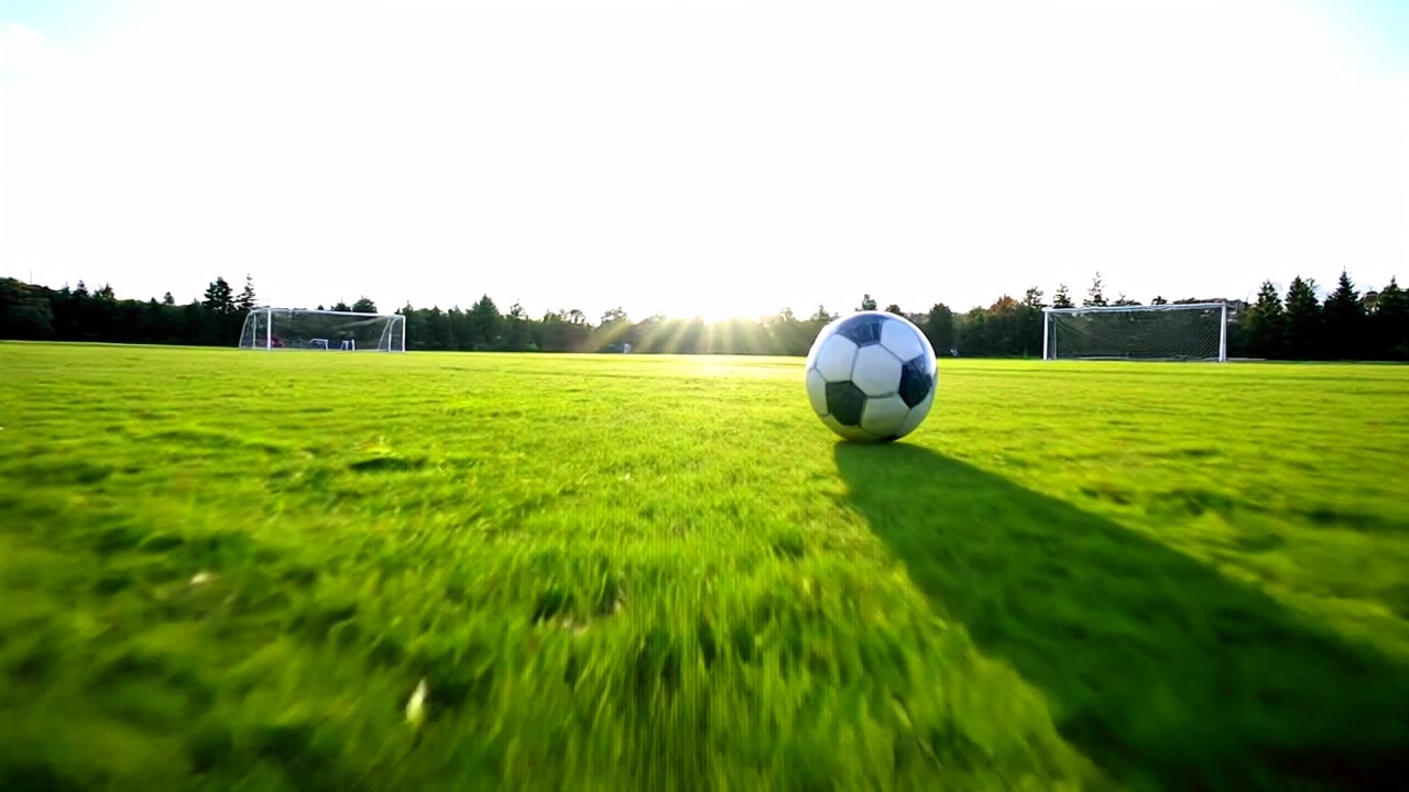 Subject: A soccer ball zipping towards a focused player. Subject movement: The ball hurtles swiftly across the field. Scene: An outdoor soccer field with green grass and goalposts in the background. Style: Bright, afternoon light illuminating the scene, creating sharp contrasts. Motion: The camera tracks the ball's rapid approach, moving in a low angle to emphasize its speed and determination.