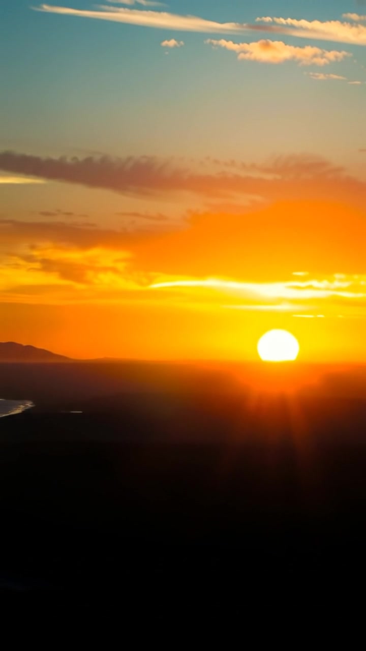 A breathtaking sunset over a distant horizon, warm golden and orange tones illuminating the sky, with soft clouds and a tranquil sea reflecting the vibrant colors. The scene is bathed in natural, gentle lighting with smooth camera movement capturing the vast landscape. The background features calming ocean waves.
