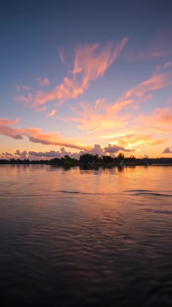A wide shot of a sunset over a tranquil lake, with vibrant colors reflected on the still water. Natural lighting bathes the scene in warm tones, with soft shadows and a smooth camera dolly shot enhancing the peaceful atmosphere. The background sound features gentle lake waves.
