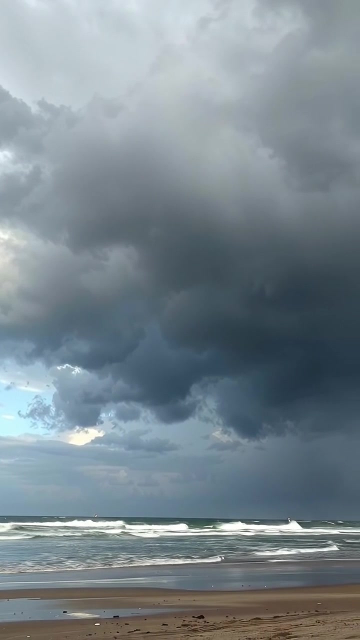 Dark storm clouds gathering over a windswept beach with swirling mist, captured in high detail with natural lighting, gentle camera pan emphasizing ominous sky and churning waves, creating a tense atmosphere. The background audio features roaring ocean waves and whipping wind.