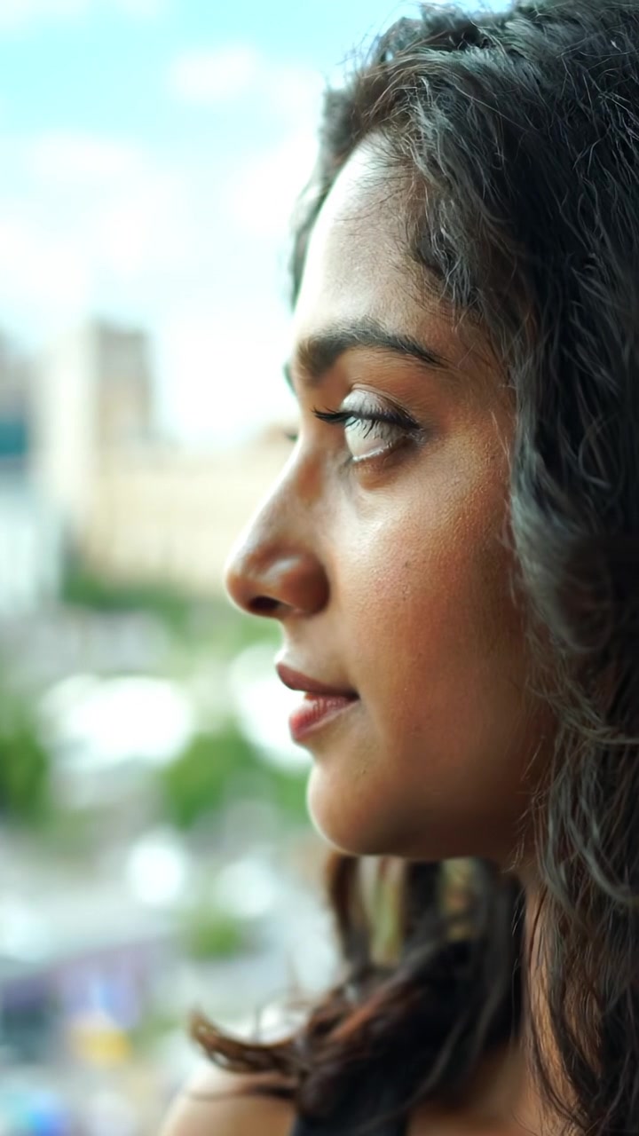 A dramatic shot of Maya gazing out of a window, lost in thought with soft natural lighting, high detail, smooth camera zoom focusing on her contemplative expression. The background features a serene cityscape under a partly cloudy sky, with gentle ambient sounds of distant city life.