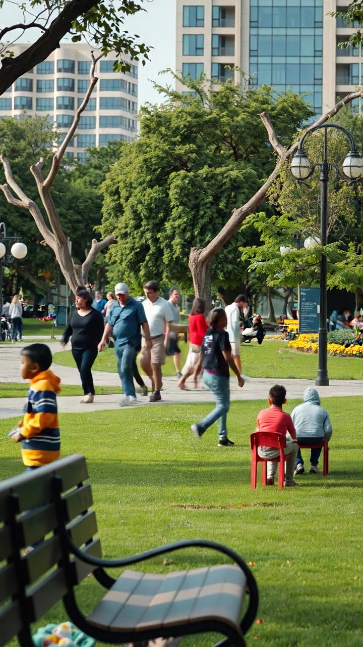 An urban park bustling with people strolling, children playing on vibrant playgrounds, and families relaxing on benches. Captured in natural light with smooth camera movements, emphasizing warmth and community spirit. The background features lush greenery and clear skies, with the sound of cheerful laughter and birdsong.