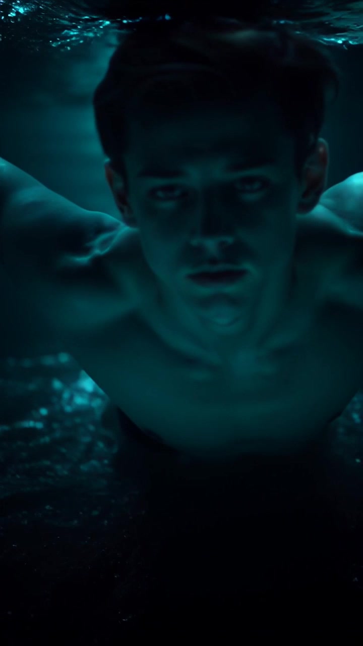 Subject: A swimmer gliding through dark waters. Subject movement: Ryan swims smoothly, kicking off from the bottom, arms pulling through the water with a rhythmic motion towards faint glimmers of light ahead. Scene: Underwater setting, surrounded by shadows and the faint sparkle of distant light. Style: Ambient lighting creating a serene yet slightly mysterious atmosphere. Motion: The camera closely follows the swimmer, panning alongside to capture the fluid movements and ripples while occasionally tilting to showcase the shimmering lights ahead.