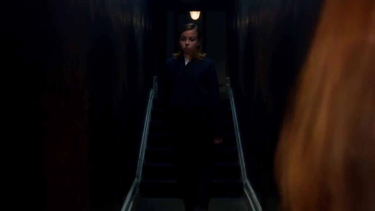 Subject: A young girl with a curious expression. Subject movement: She steps cautiously forward, glancing up the dark staircase leading to the attic. Scene: A dimly lit hallway dominated by a steep, looming staircase, shadows playing across the walls. Style: Soft, ambient lighting with an emphasis on contrasts between light and dark, evoking a sense of mystery. Motion: The camera gently pushes in on her as she gazes up, creating a sense of anticipation and intrigue.