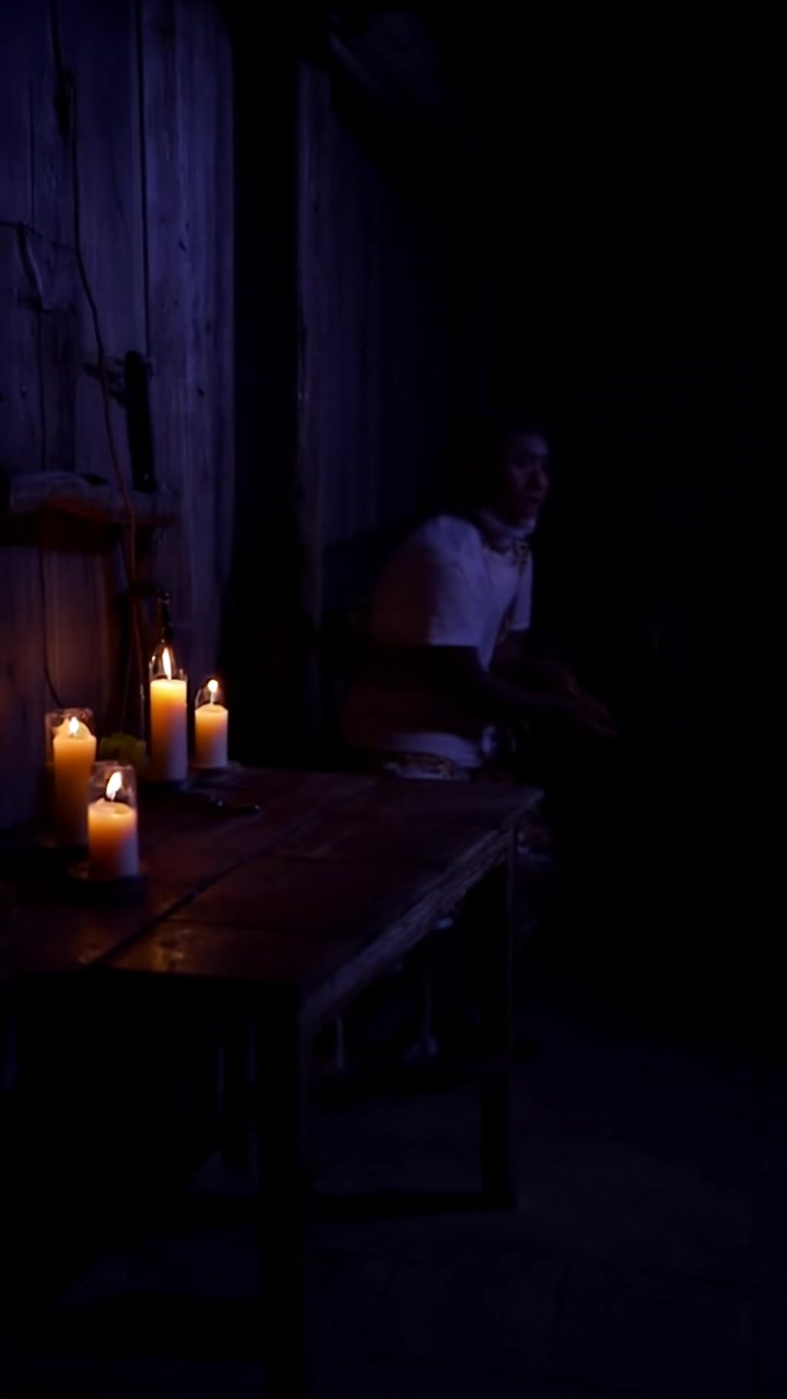 Subject: A person standing in a dimly lit room, looking around nervously with a worried expression. Scene: The intimate, shadowy space is adorned with faintly flickering candles and rustic wooden furniture. Style: Mysterious ambiance enhanced by soft, flickering candlelight casting dancing shadows. Motion: The camera subtly tracks in towards the subject, capturing their anxious movements and expressions as they shift from side to side, heightening the tension in the scene.