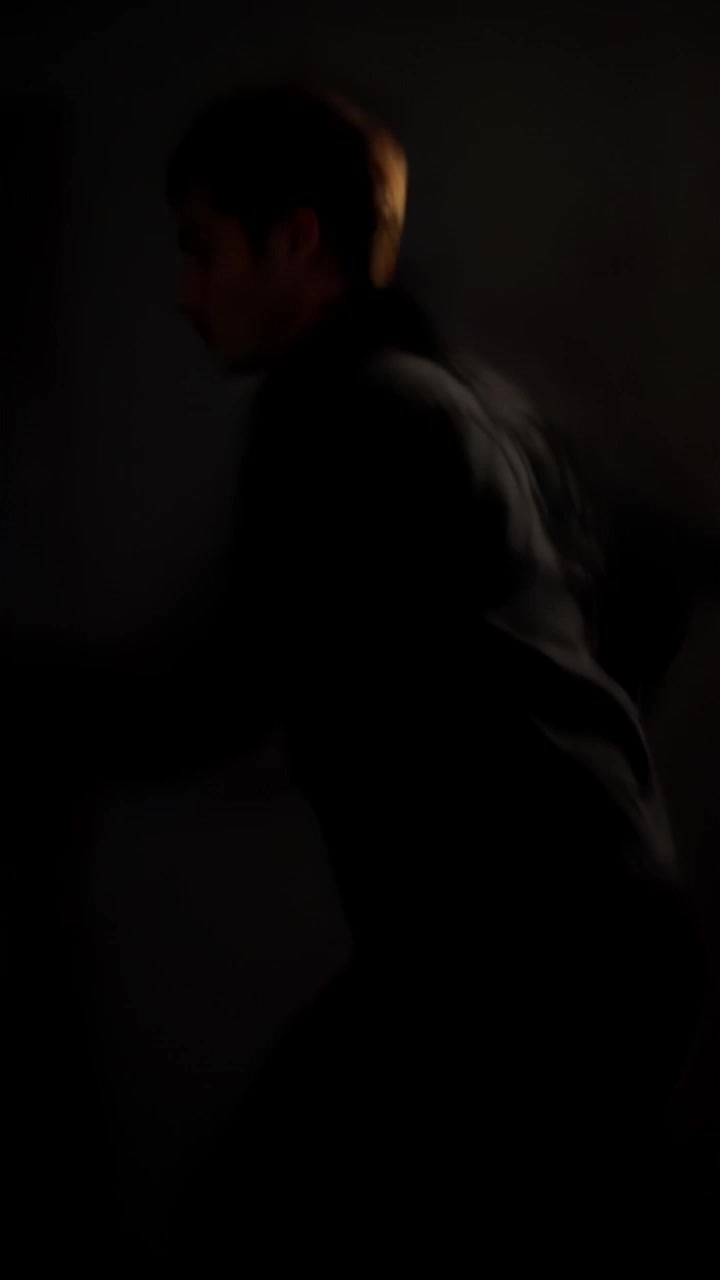 Subject: A person running through a dimly lit hallway with a look of urgency. Subject movement: The person dashes swiftly, taking long strides as they navigate the corridor. Scene: A narrow, shadowy hallway with flickering lights casting dramatic shadows. Style: Low-key lighting creating a tense atmosphere, accentuating the urgency of the moment. Motion: The camera trails closely behind the person, capturing their hurried movements while dynamically shifting angles to enhance the sense of speed and urgency.