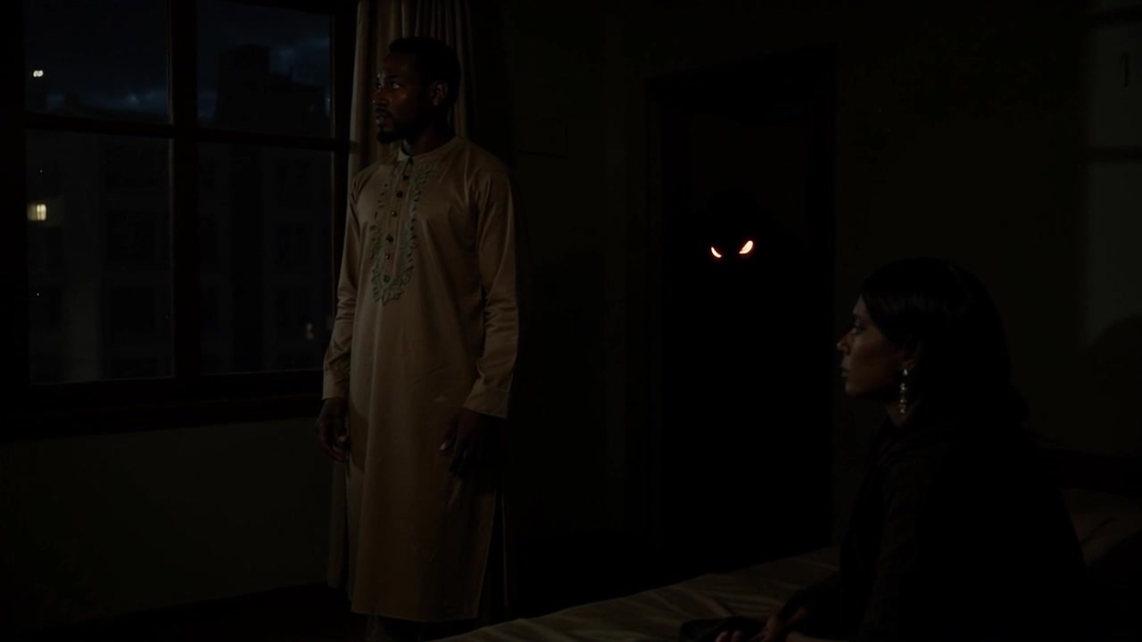 Scene: A dimly lit room with flickering shadows, featuring a man in traditional attire standing near a window and a woman sitting on a bed, all while a shadowy figure with glowing eyes looms in the doorway behind them. Style: Atmospheric lighting with deep shadows, creating a suspenseful and mysterious ambiance. Motion: The camera slowly dolly-in toward the couple, heightening tension as it subtly reveals the ominous figure watching from the shadows.