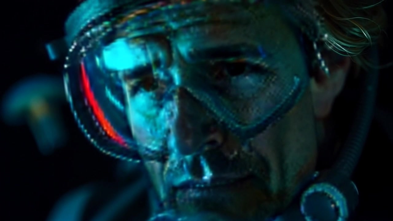 Scene: A close-up of a diving mask reflecting the face of an individual, showing visible strain and fear. Style: Dim underwater lighting accentuates the tension, creating a suspenseful atmosphere. Motion: The camera slowly pulls back from the diving mask, revealing a vast, murky underwater environment lurking in the background.