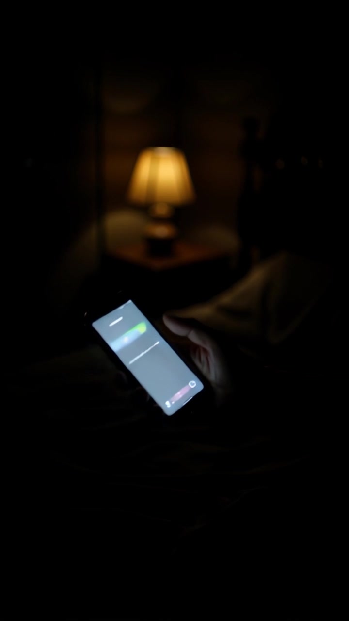 Scene: A dimly lit bedroom, softly illuminated by a bedside lamp, creating a cozy, intimate atmosphere. Motion: The camera gently pushes in on the hand holding the smartphone, showcasing the incoming call notification from 'DEBBIE KP'. Style: Warm, muted lighting enhances the room's tranquility as the camera encroaches on the phone, inviting the viewer into the moment.