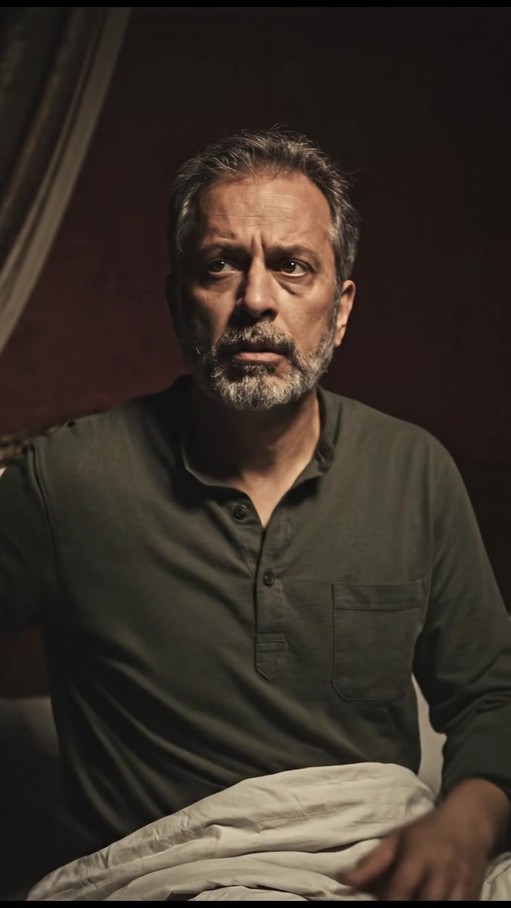 Raj, in a dimly lit bedroom, sits up in bed, looking confused with shadows around him.